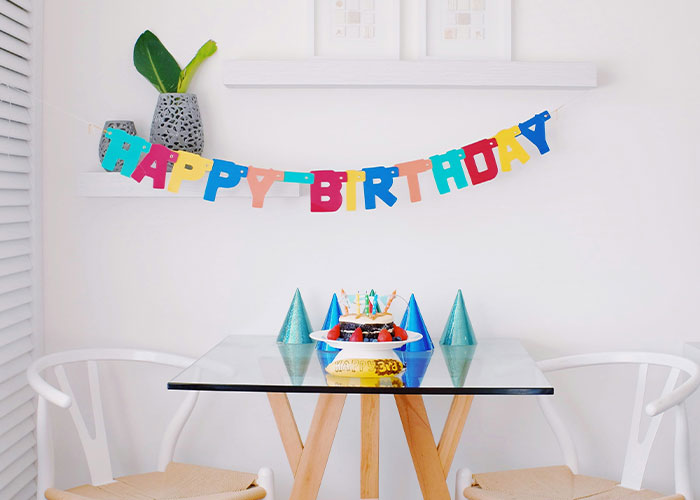 Colorful happy birthday banner above a small table with party hats and a cake, reflecting strange childhood realities.