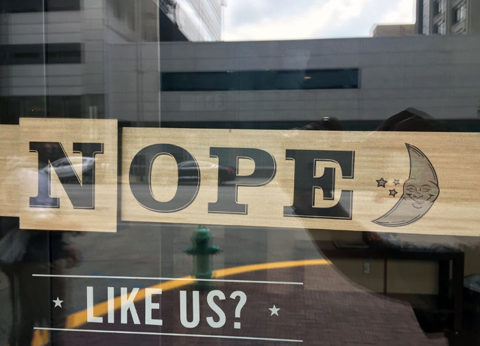 Sign on a bar or restaurant window with the word NOPE and a crescent moon graphic reflecting a city street scene.