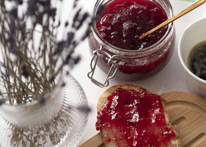 Jar of red jam with spoon, toast spread with jam, coffee cup, and blurred lavender in a vase on a table.