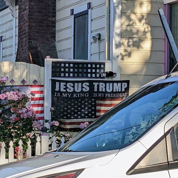 Sign fail with text "Jesus is my king, Trump is my president" on a house fence next to flowers.