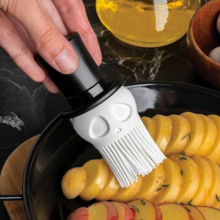 Weird kitchen item: skull-shaped basting brush applying oil to sliced potatoes.