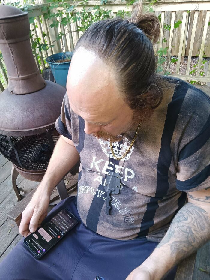Man with a partially bald head and long hair, using a phone on a wooden deck; example of a hair accident.