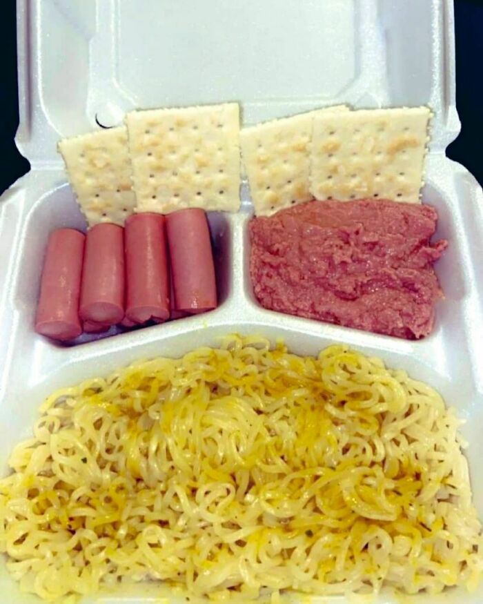 Unappetizing meal with instant noodles, crackers, sausages, and pink meat in a styrofoam container.
