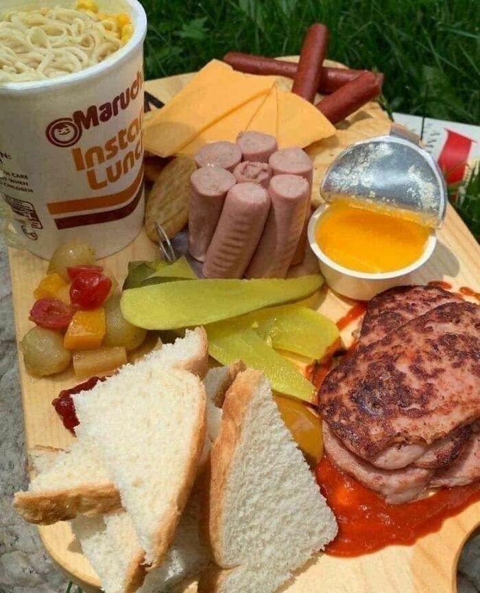 Unappetizing food display with instant noodles, processed meats, cheese slices, bread, and pickles on a wooden board.