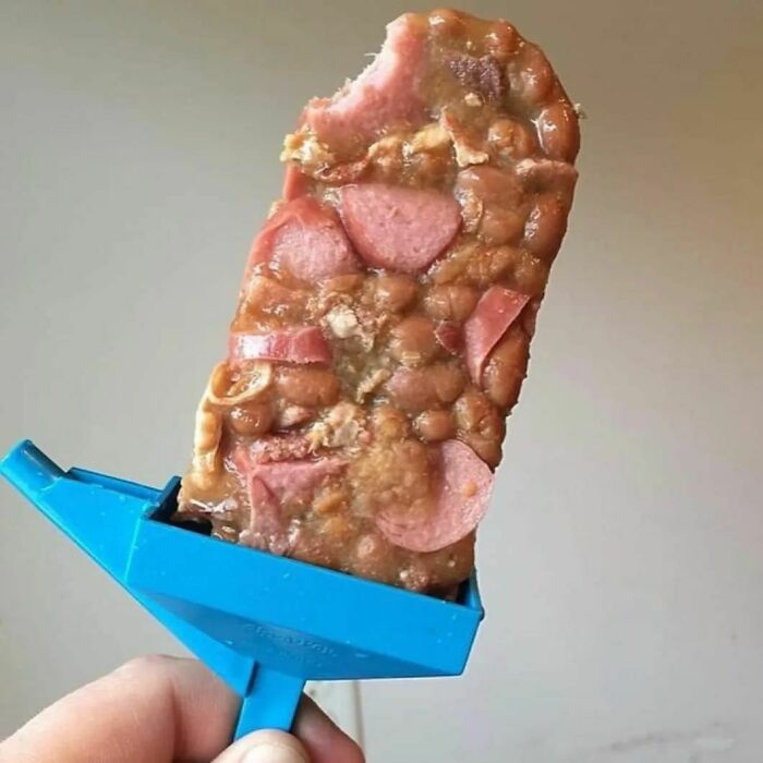 Unappetizing food: beans and hot dogs on a stick with a blue handle, held against a neutral background.