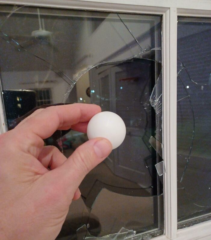 Hand holding a ping pong ball in front of a window with a large shattered glass pane showing parents having a miserable day.