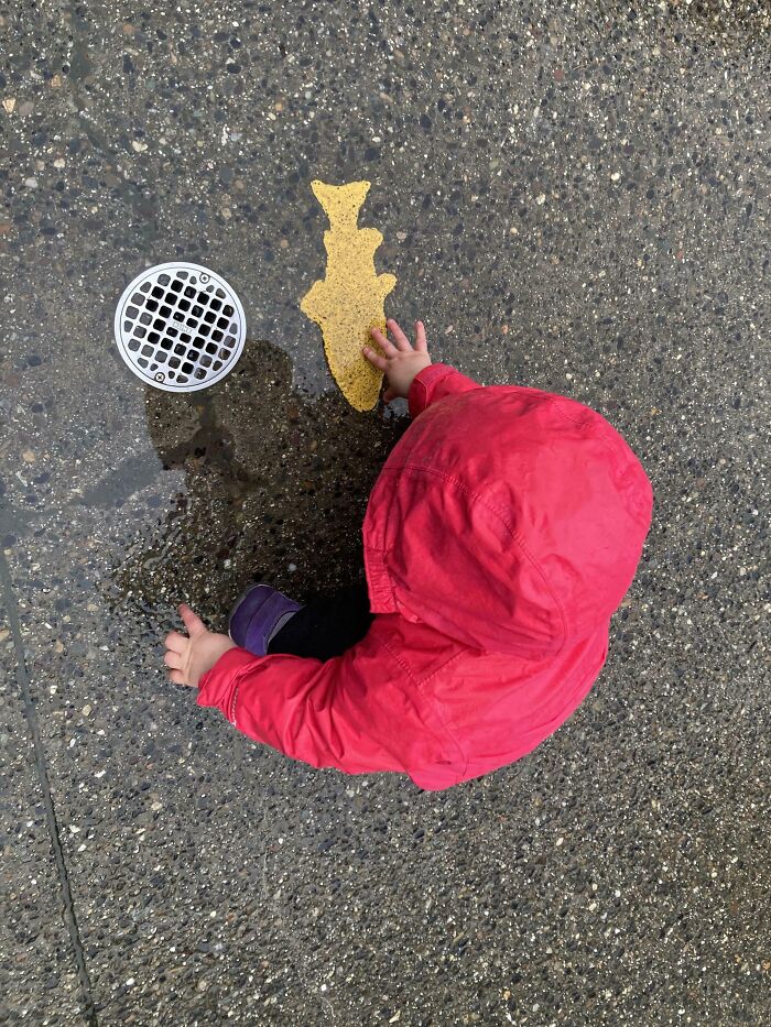 Toddler in red rain jacket playing on wet pavement near a drain, capturing a moment reflecting parents having a miserable day.