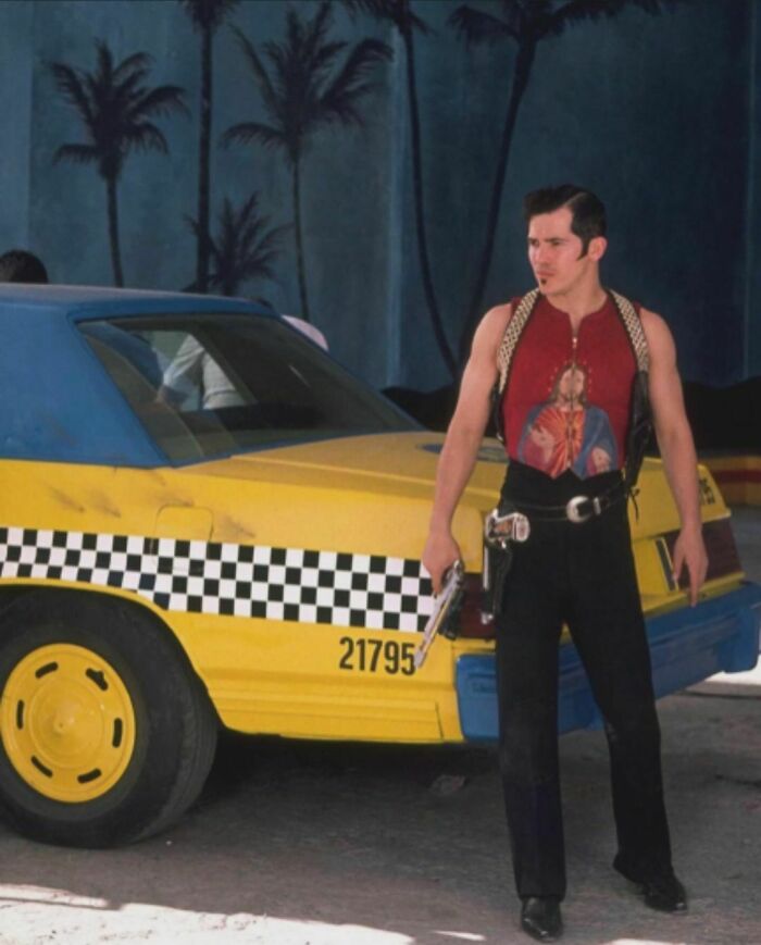 Man in 90s fashion stands by a yellow taxi, wearing a red graphic tank top and black pants, evoking nineties nostalgia.
