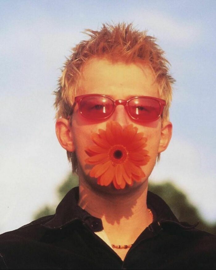 Person with spiky hair and red sunglasses holding an orange flower to their face, encapsulating nineties nostalgia.