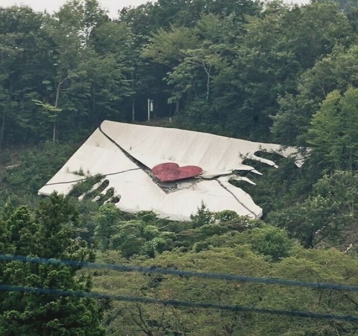 Oversized hands revealing a heart on a mountainside, symbolizing 90s creativity amidst lush greenery.