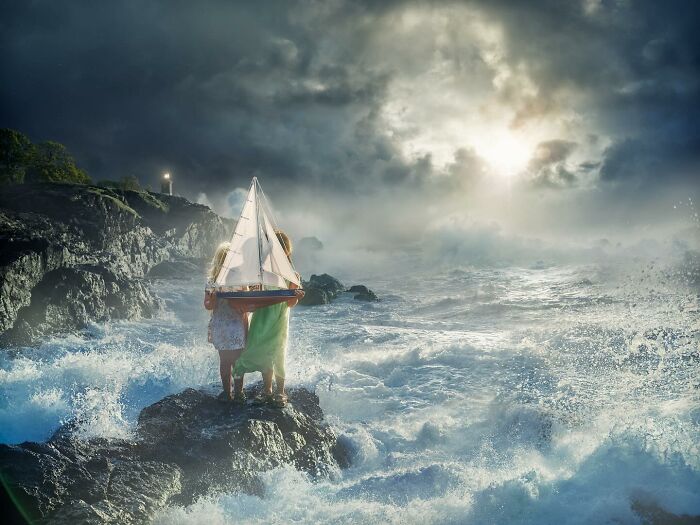 Two people holding a sailboat on rocky shore amid stormy seas, showcasing mind-bending photo manipulation.