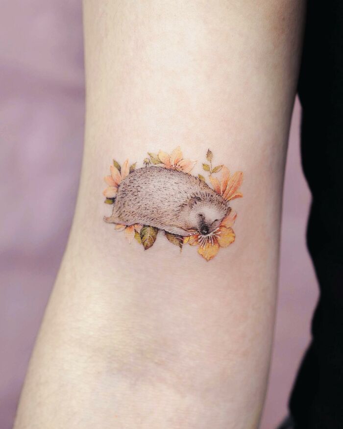 Realistic tattoo of a hedgehog surrounded by flowers on an arm.