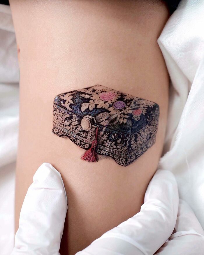 Realistic tattoo of an ornate box on skin, created by a talented artist, showcasing intricate floral designs.