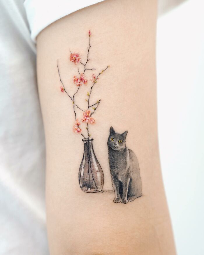 Realistic tattoo of a black cat sitting beside a vase with pink blossoms.