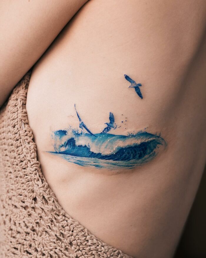 Realistic tattoo of a wave and bird in blue ink on skin.