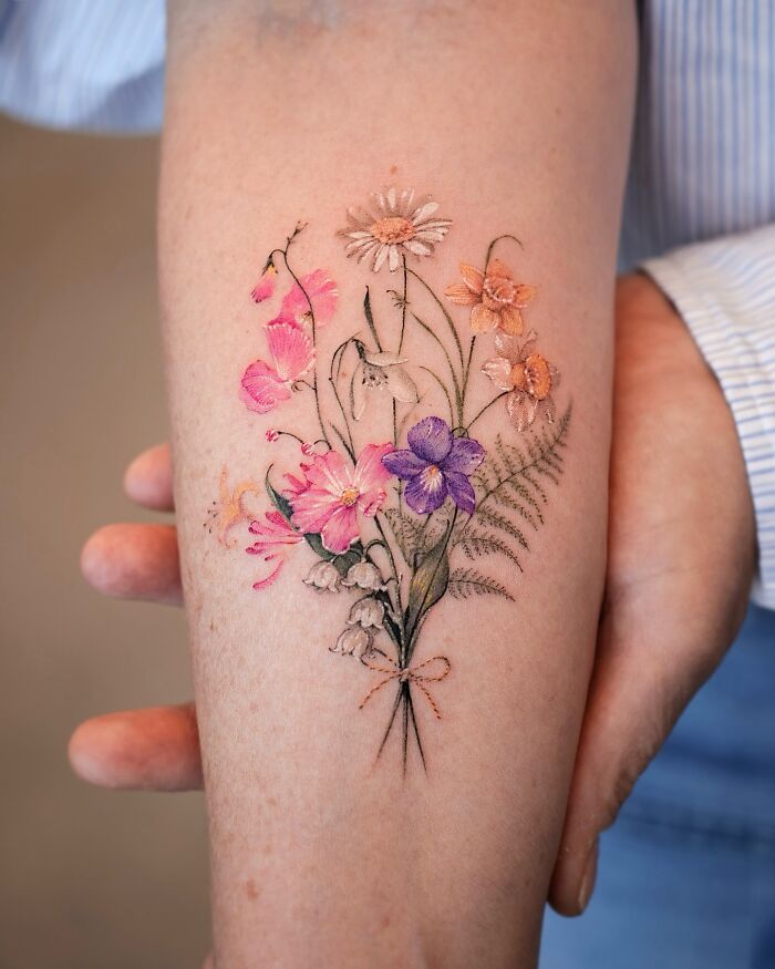 Realistic floral tattoo on a forearm, showcasing vibrant pink, purple, and orange flowers with green leaves.
