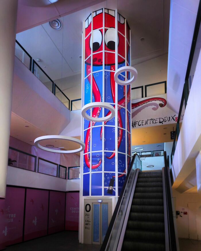 Humorous street art by Oakoak depicting a playful octopus around a glass column in a shopping center.