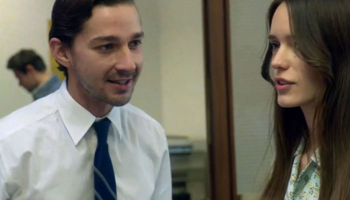 Movie stars in a film scene, wearing office attire and engaging in conversation in an intimate setting.