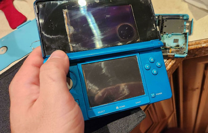 Hand holding a broken blue handheld gaming console with parts detached, illustrating parents having a pretty miserable day.