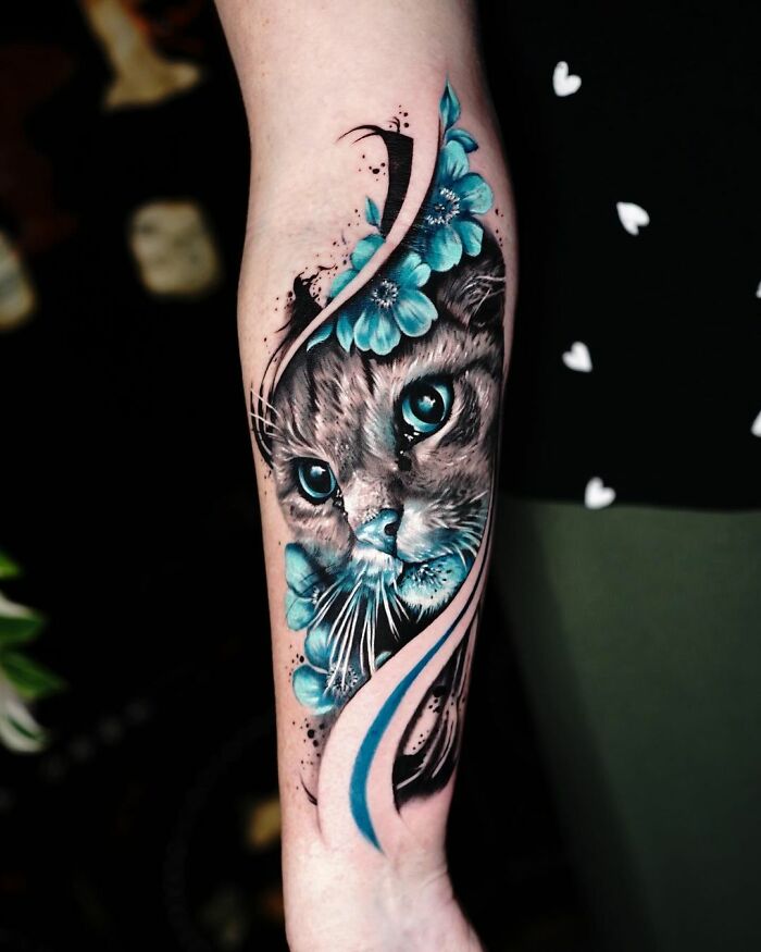 Cute pet portrait tattoo featuring a realistic cat with blue eyes and floral elements on a person's forearm.