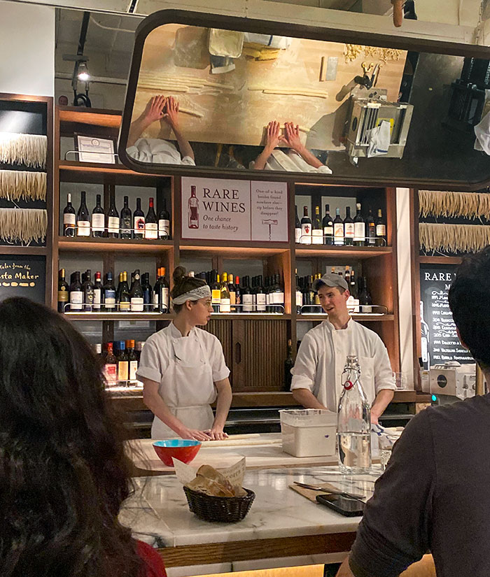 Two chefs preparing food behind the bar in a restaurant with bottles of wine in the background.