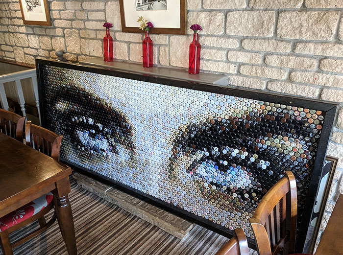 Mosaic artwork of eyes made from bottle caps displayed in a bar setting with wooden chairs and stone walls.