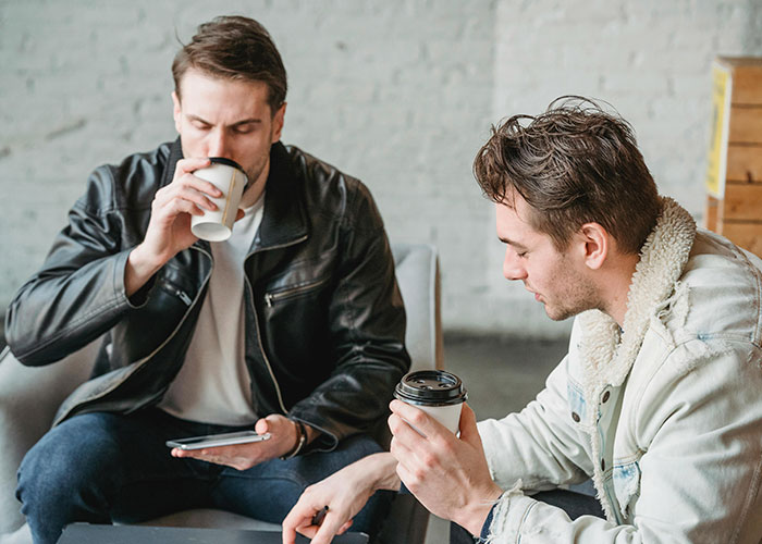 Two men in jackets sipping coffee, discussing cringe-worthy experiences in a casual setting.