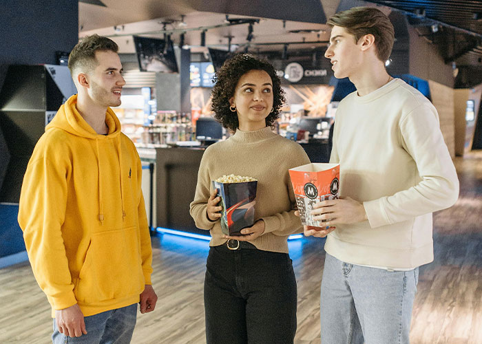 Three people at a movie theater, holding popcorn, standing and talking indoors.