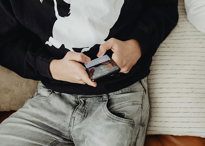 Person in a black sweatshirt and jeans using a smartphone, possibly recalling a cringe-worthy experience.