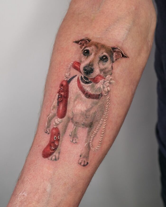 Tattoo of a cute pet portrait featuring a dog holding a sausage and rope toy on an arm.
