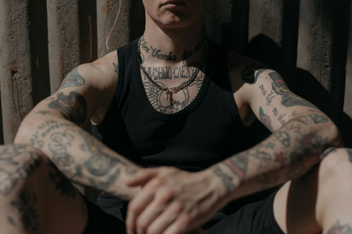 Young man with tattoos on arms and neck, wearing a black tank top, sitting against a corrugated metal wall.