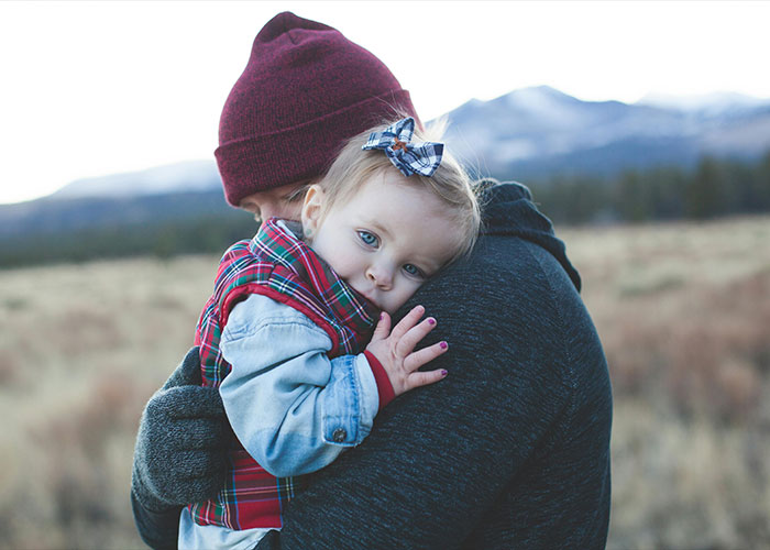 Adult wearing a red beanie holding a toddler in a plaid jacket, illustrating strange childhood realities and emotional bonds outdoors.