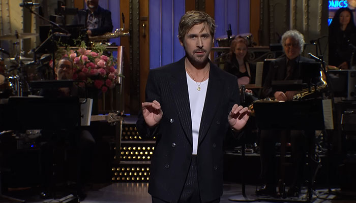 Actor on stage in a dark suit, hands gesturing, with a band and floral arrangement in the background.