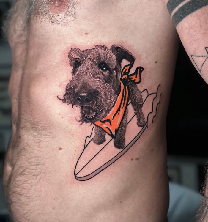 Tattoo of a dog with an orange bandana riding a surfboard, merging two artistic styles seamlessly.