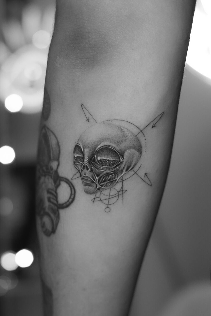 Single needle tattoo of an alien with geometric patterns on forearm, showcasing intricate design and shading.
