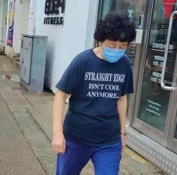 Person wearing a mask and a cringe shirt walking on the sidewalk outside a fitness center in a random humorous photo.