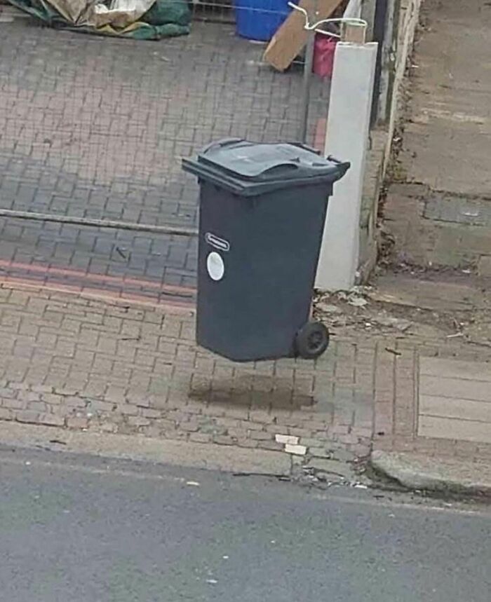 Black trash bin appears to be floating above pavement in a random and cringe photo with unique humor appeal.
