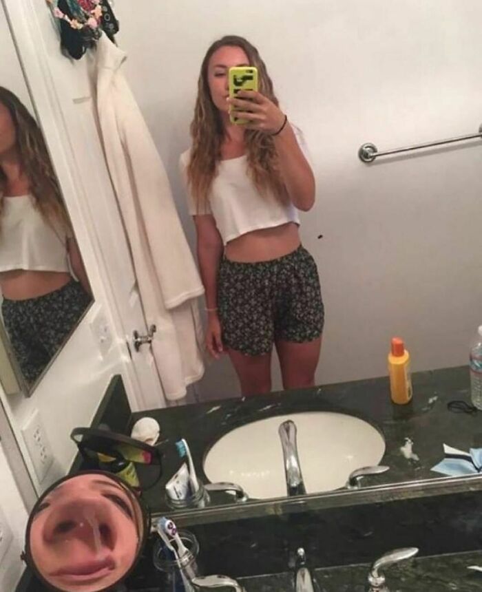 Young woman taking a bathroom mirror selfie with a funny distorted nose reflection in a random and cringe photo.