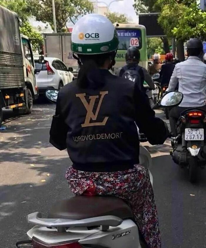 Person on scooter wearing a jacket with a parody logo and text, captured in a random and cringe photo on a busy street.