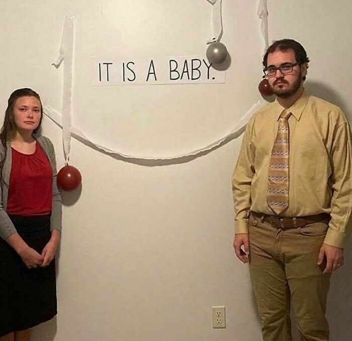 Two people standing beside a wall with a sign reading "IT IS A BABY" in a random and cringe photo with unique humor.