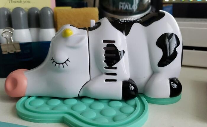A quirky cow-shaped device with black spots and closed eyes, resting on a green stand among various desk items.