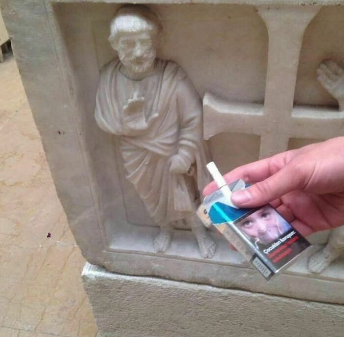 Hand holding cigarette pack near ancient statue, capturing a bizarre moment from a weird Instagram account.