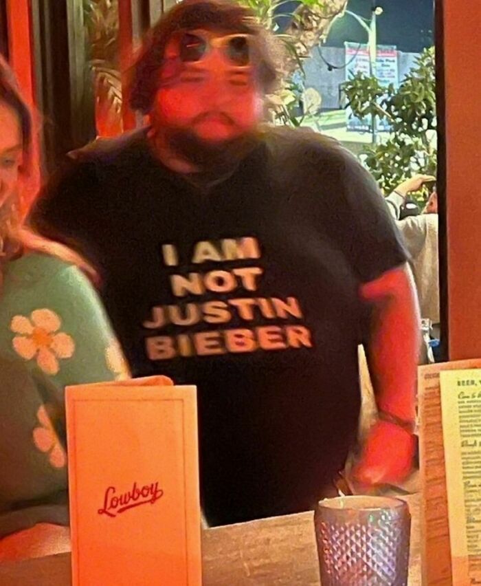 Man at a bar in a "I am not Justin Bieber" shirt, embodying the quirky style of the "I Want To Leave Ok" Instagram account.