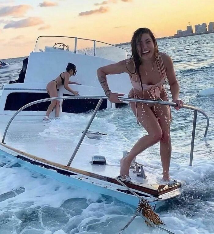 Women balancing on a sinking boat deck, laughing amidst ocean waves at sunset.