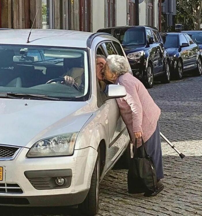 Elderly woman kisses man through car window on a city street, showcasing the quirky charm of I Want To Leave Ok.