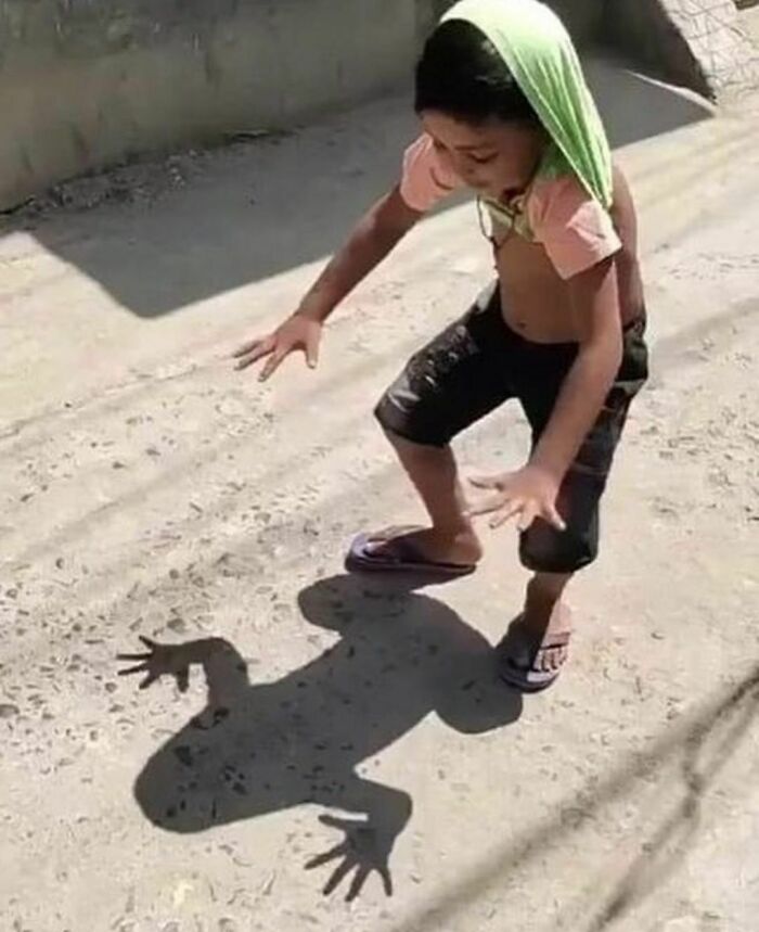 Child with a shirt on their head and hands raised casts a lizard-like shadow on the ground in a funny Instagram pose.