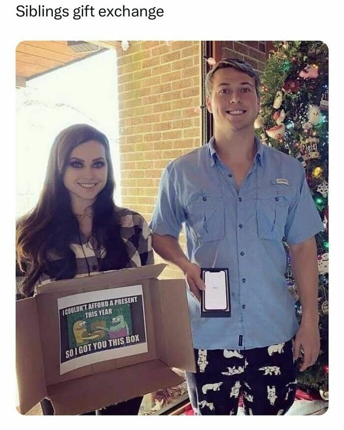 Siblings exchanging funny gifts by a Christmas tree; one holds a box with meme, the other a smartphone box.