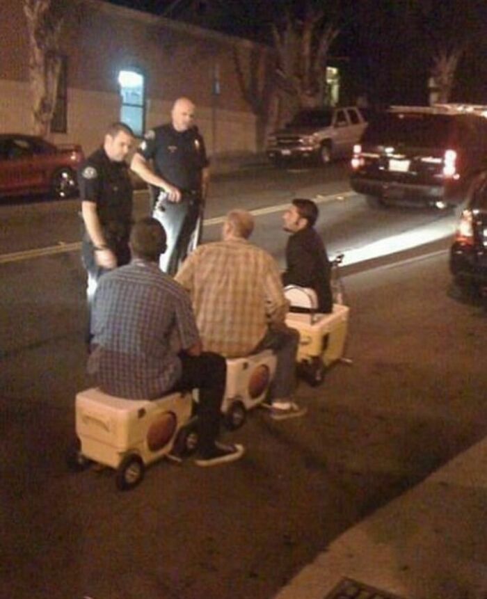 Three men sitting on coolers like toy cars while two police officers talk to them on a street, awkward photos moment at night.