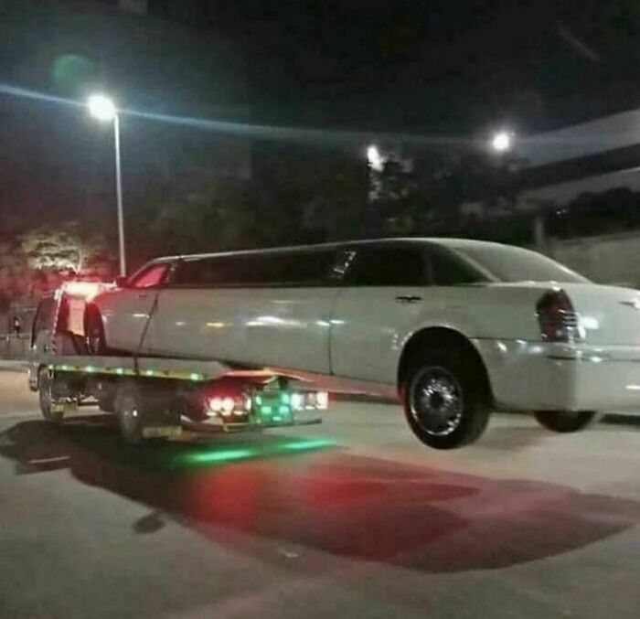 White limousine awkwardly being towed at night on a flatbed truck, capturing an awkward photo on a city street.