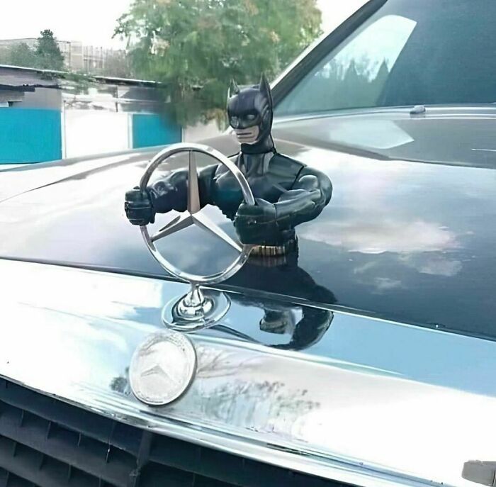 Batman figure awkwardly holding a Mercedes hood ornament on a car, creating an awkward photo moment.